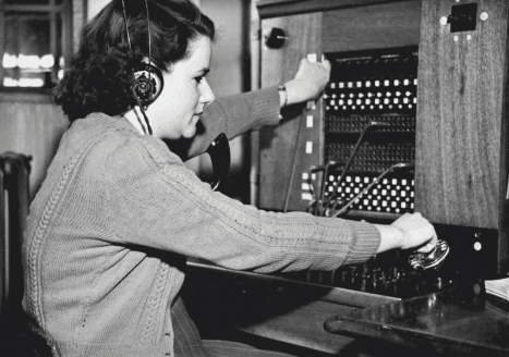 A vintage black and white photograph of a female telephone operator wearing a headset and working at a manual switchboard.