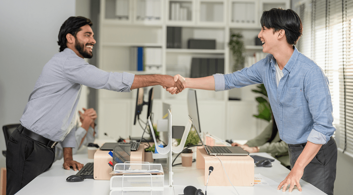 Two smiling men, wearing shirts, shake hands across a desk in a modern office