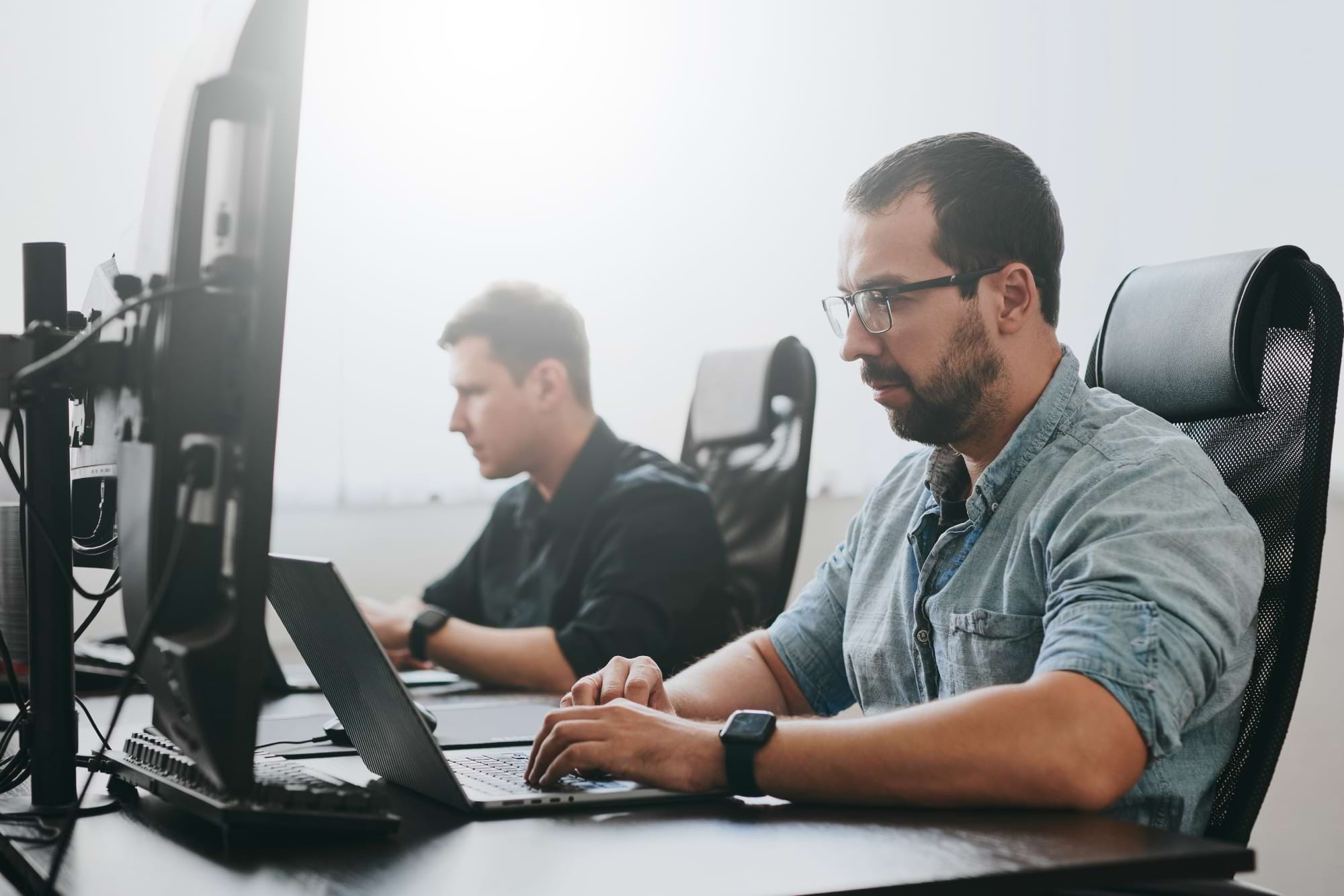 Two men are sitting in ergonomic chairs and working at their computers in a bright office space