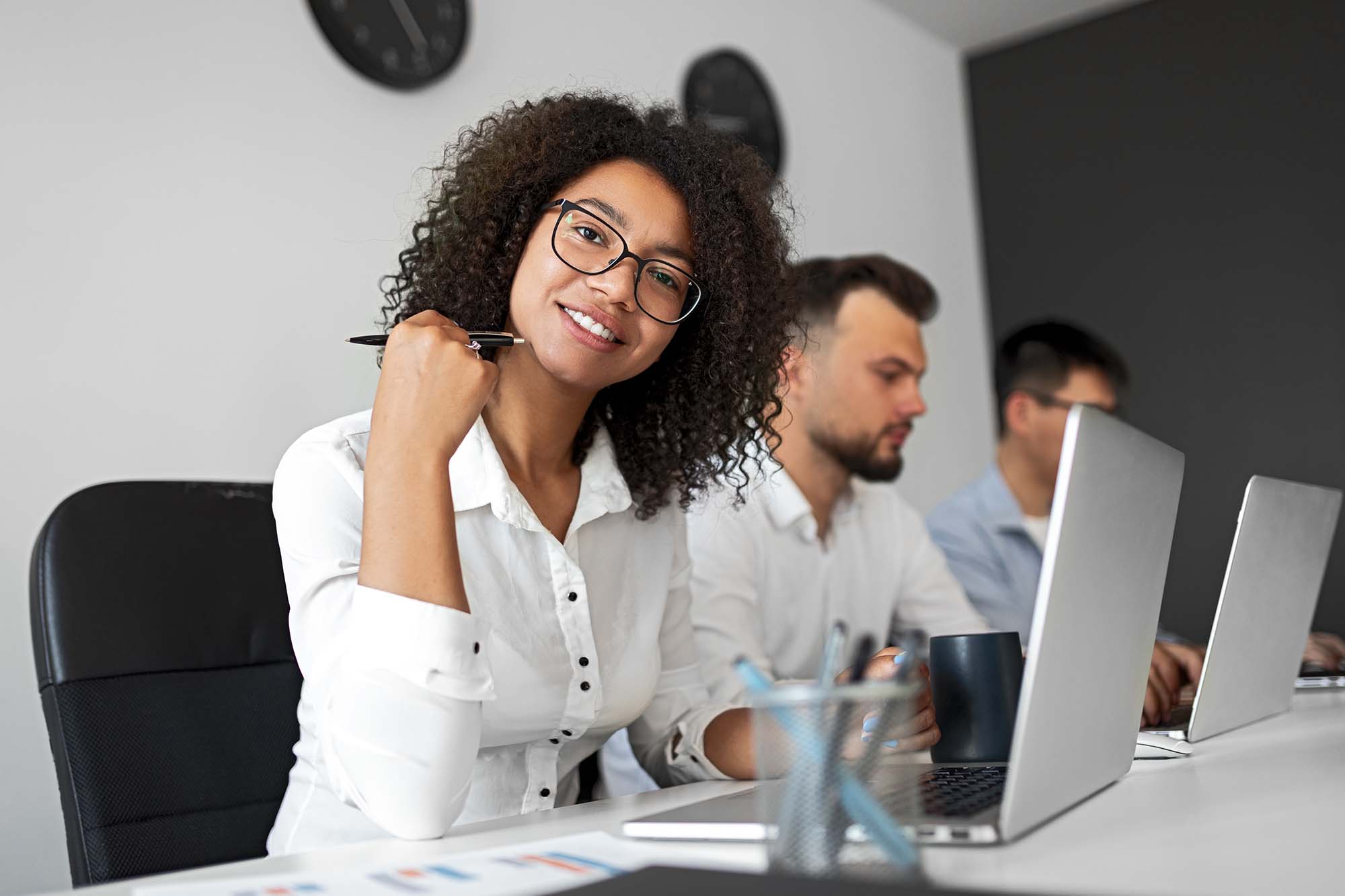 Woman with curly hair and glasses smiling at a desk with colleagues in the background.