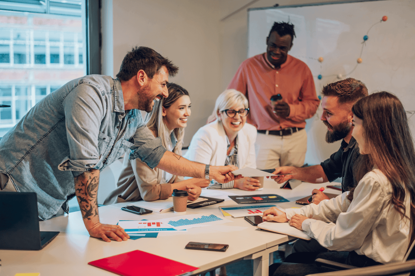 A group of professionals is gathered around a table in a lively meeting, exchanging documents and smiling.