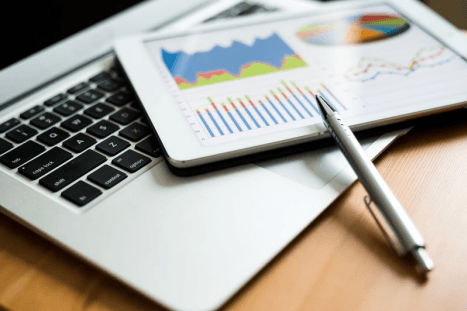 Close-up of a laptop, a tablet displaying business graphs and charts, and a pen on a wooden surface.
