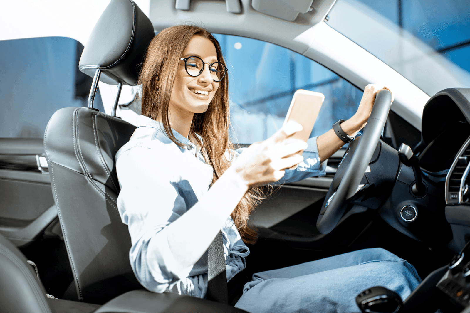 A smiling woman with glasses is sitting in a car, holding and looking at her smartphone.
