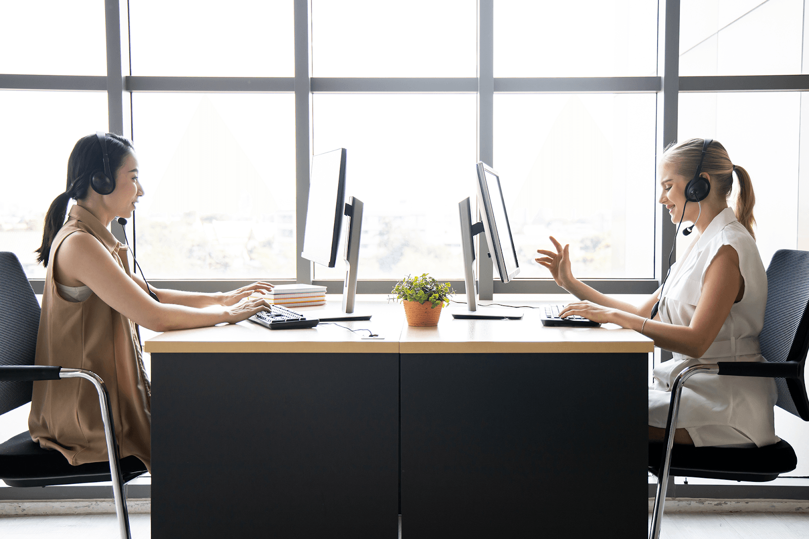 Two women, wearing sleeveless t-shirts and headsets, are sitting opposite each other and working at the computers