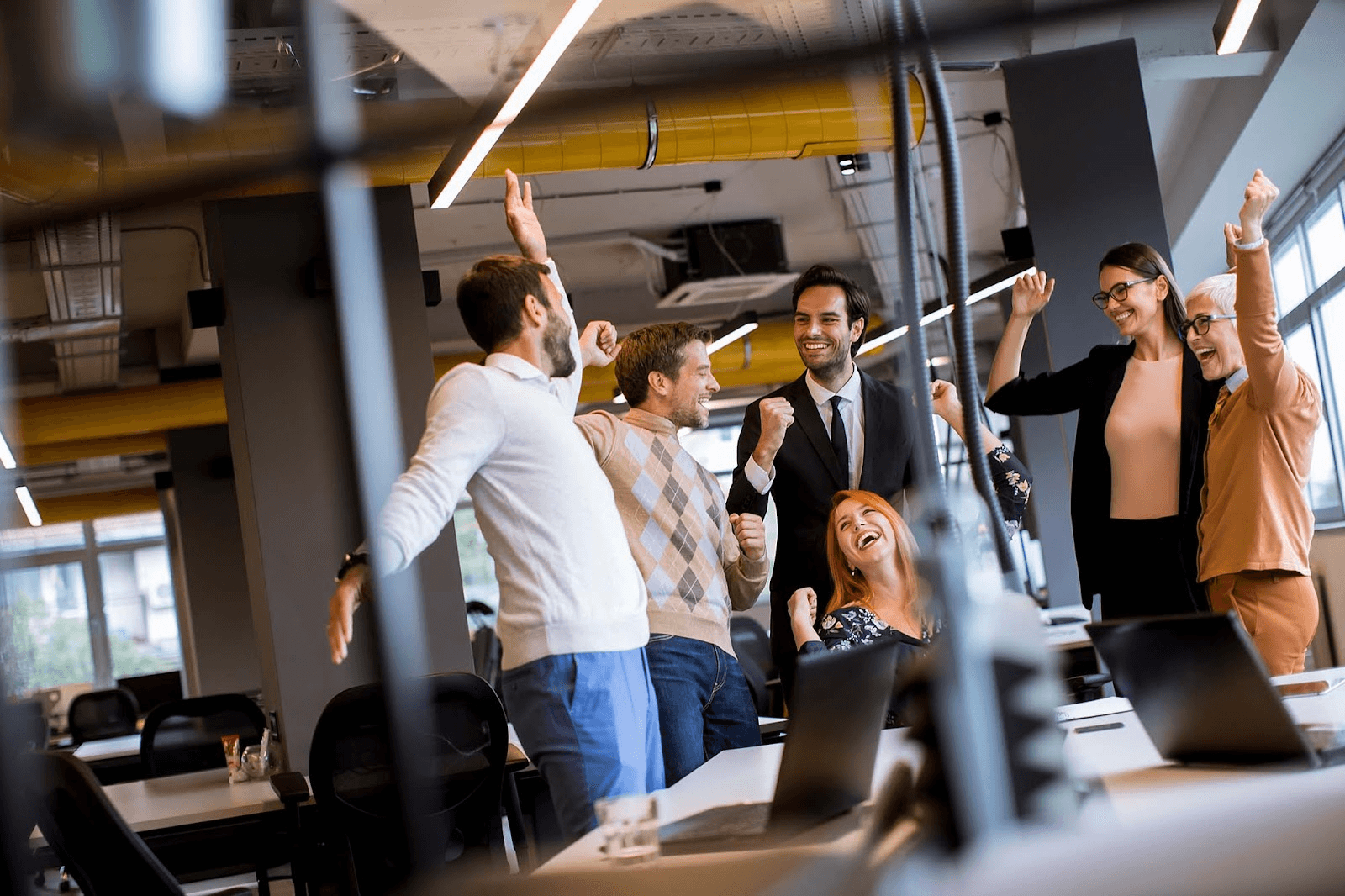 A group of business people is celebrating in an office, raising their arms in the air and smiling.