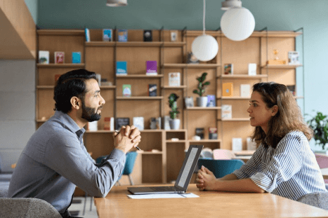 A man and a woman are sitting across from each other at a table with a laptop, engaged in a conversation.
