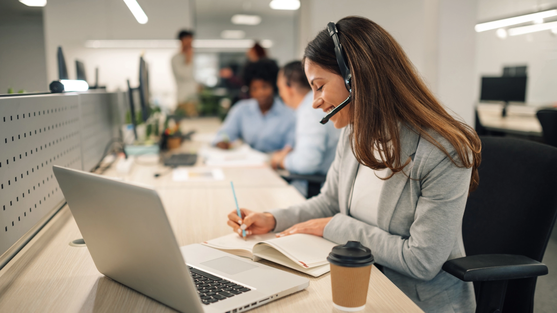 Smiling customer service representative with headset taking notes while working on a laptop.
