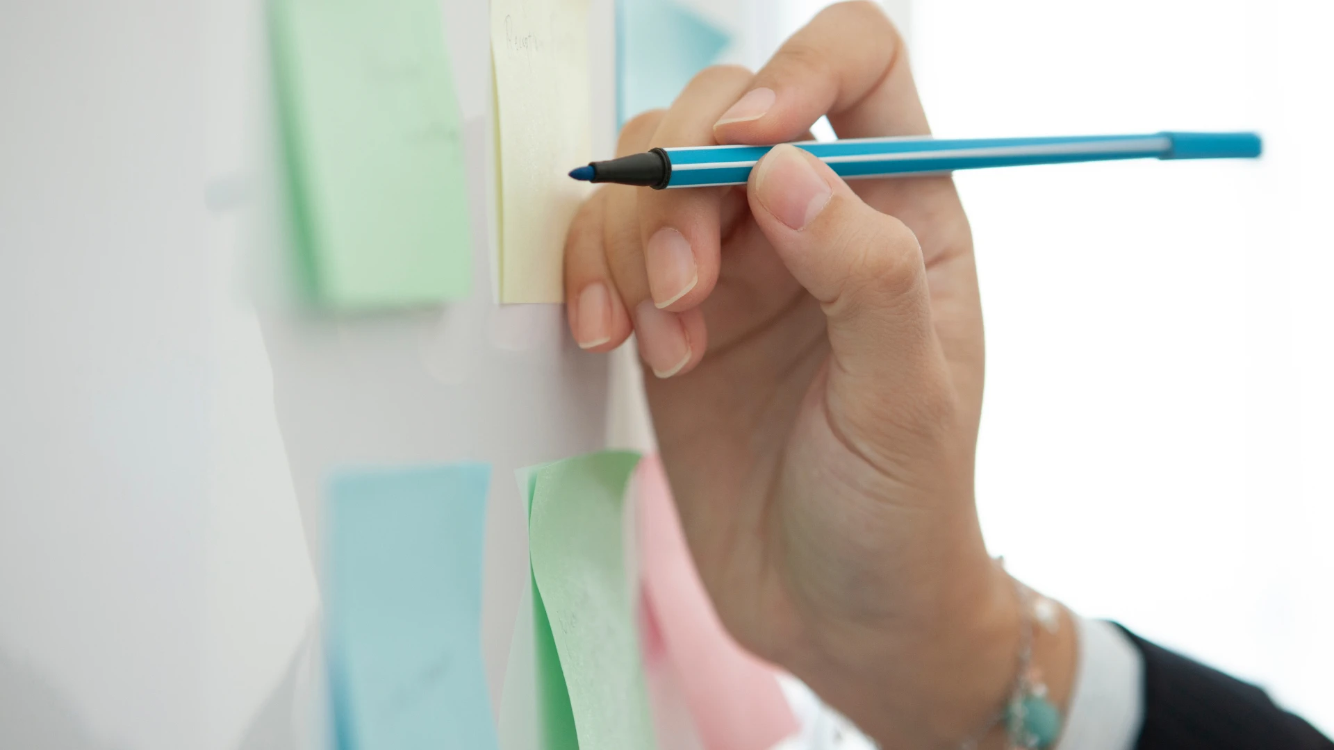 A hand holding a blue pen and writing on a yellow sticky note.