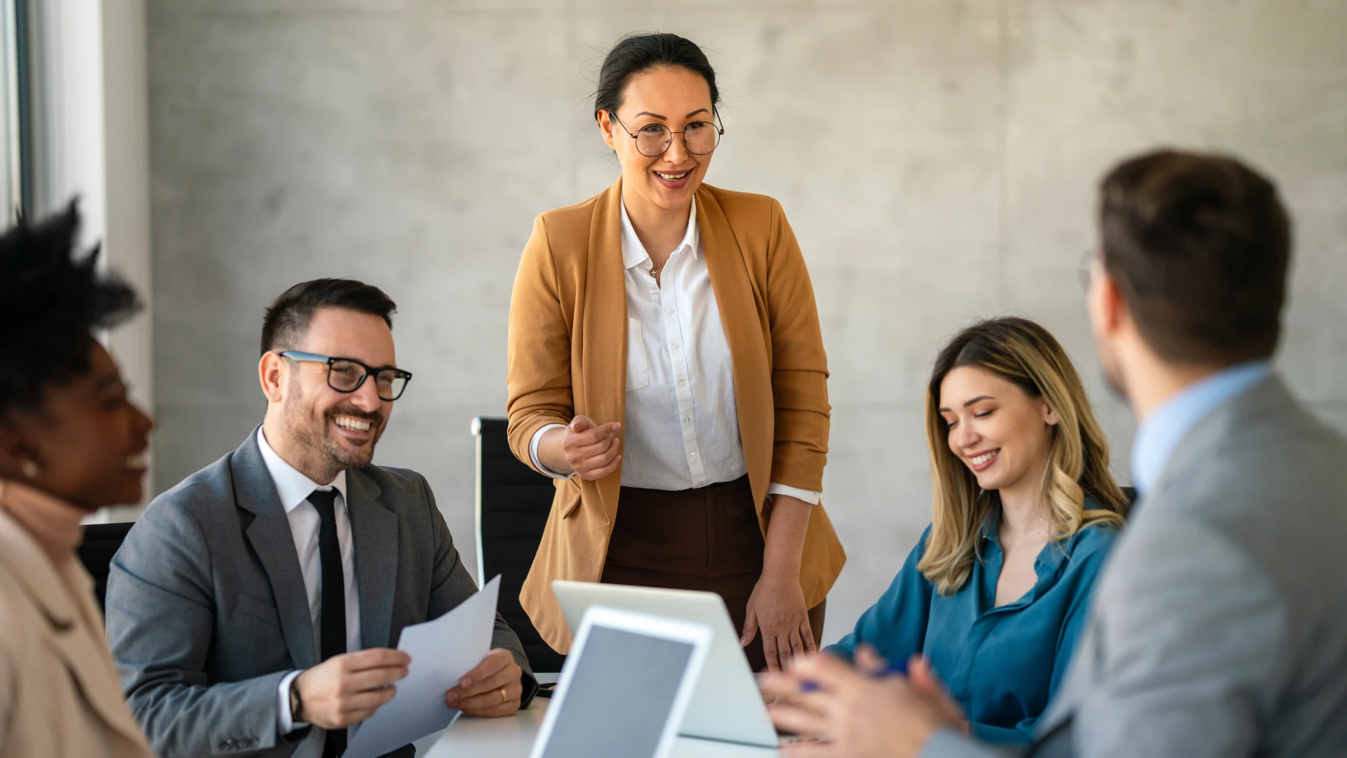 A diverse group of five professionals in a modern office having a meeting. A woman in a tan blazer is standing and speaking confidently while others are seated, smiling and listening. Laptops and documents are on the table, indicating a collaborative business discussion.