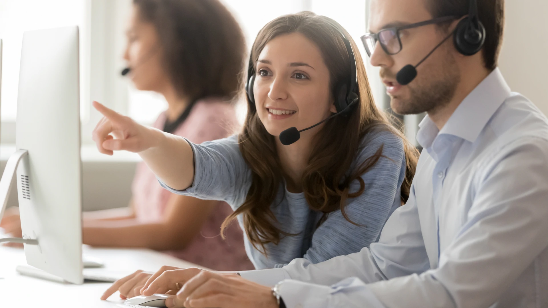 Two customer service representatives wearing headsets and looking at a computer screen. The woman is pointing to the screen.
