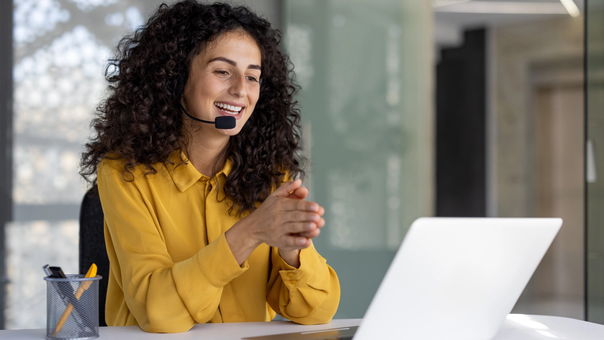 Smiling woman with curly hair wearing a yellow shirt and a headset, working on a laptop at a desk.