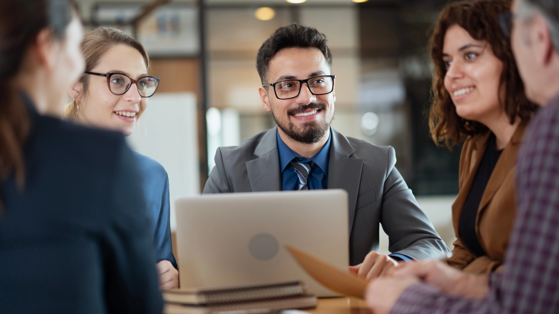 Group of business professionals in a meeting, with a man in a grey suit smiling and looking at the group.