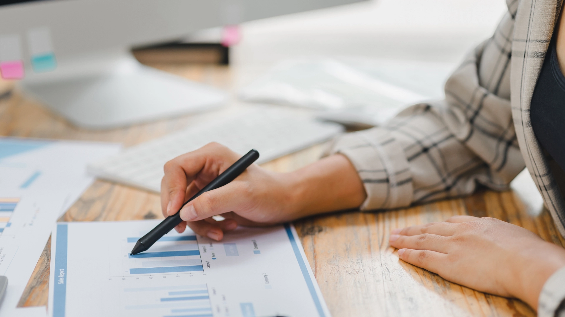 Close-up of hands using a pen to analyze charts and graphs at a desk with a computer.