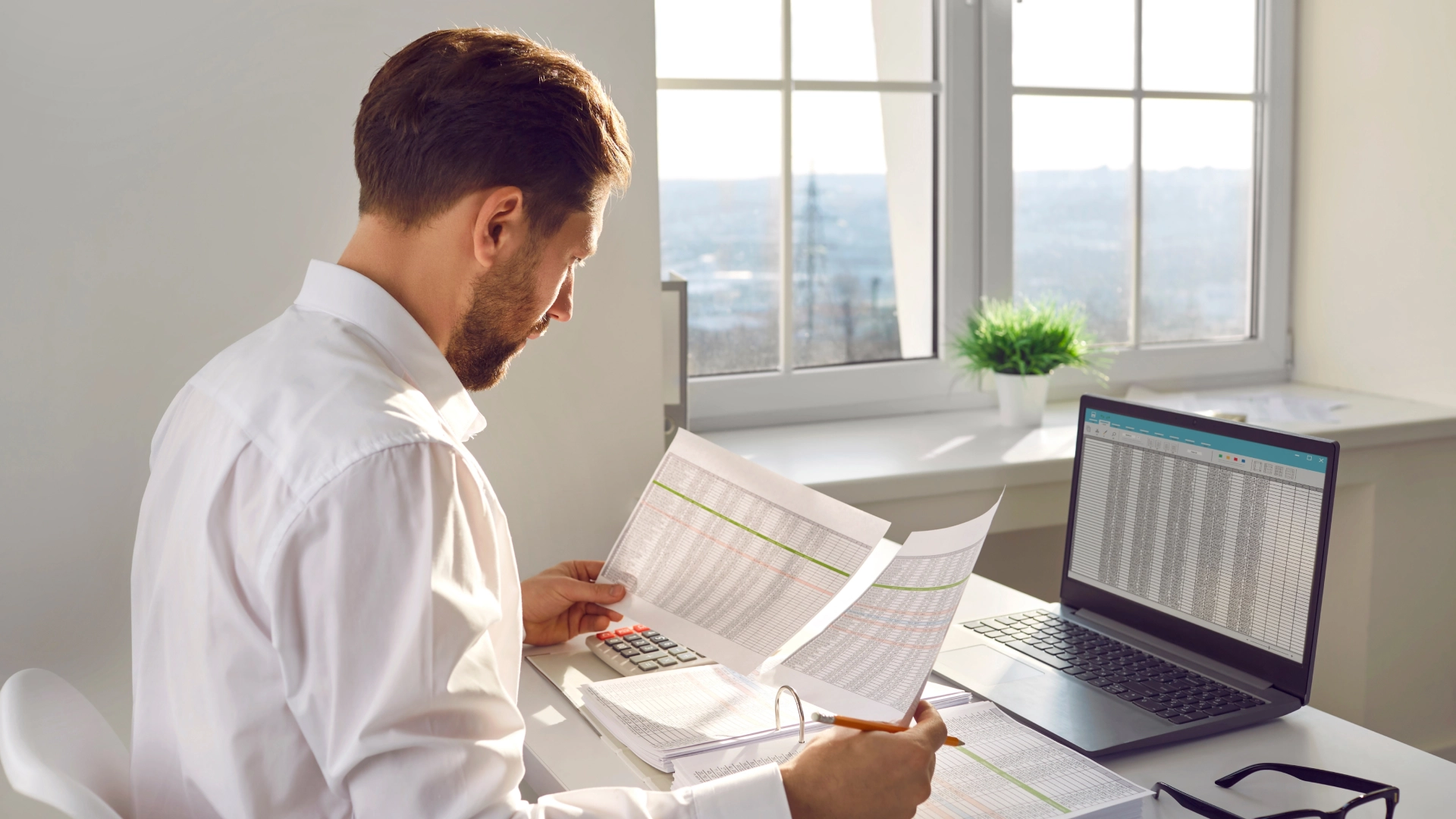 At his workstation, a man in a white shirt is closely analyzing data, reviewing spreadsheets, and using a laptop, calculator, and pencil.