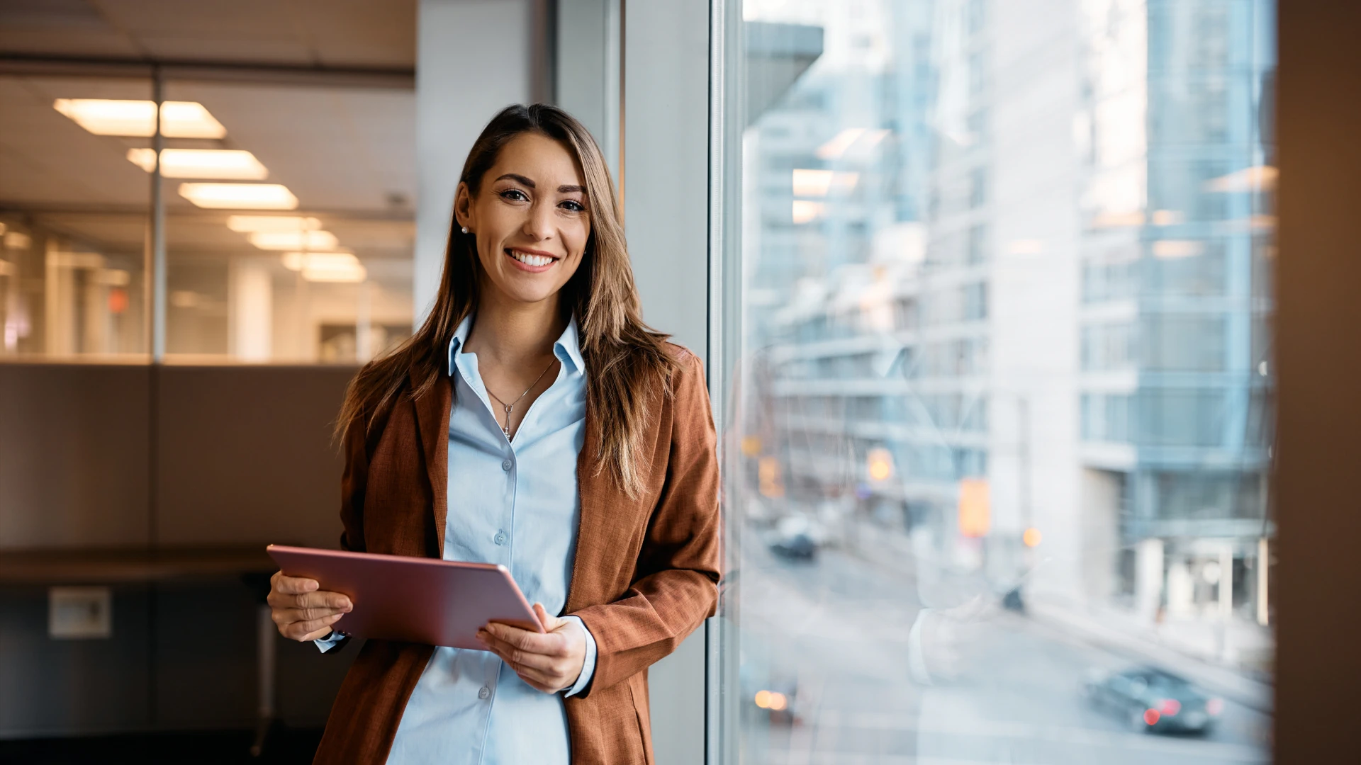 Smiling businesswoman wearing a brown blazer and light blue shirt, holding a pink tablet in front of a window in a modern office.