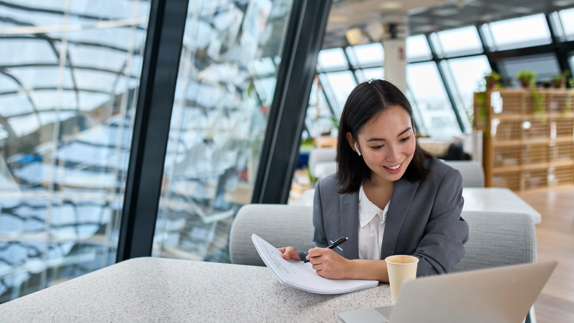 Smiling woman wearing a gray blazer and airpods, writing in a notebook at a table.