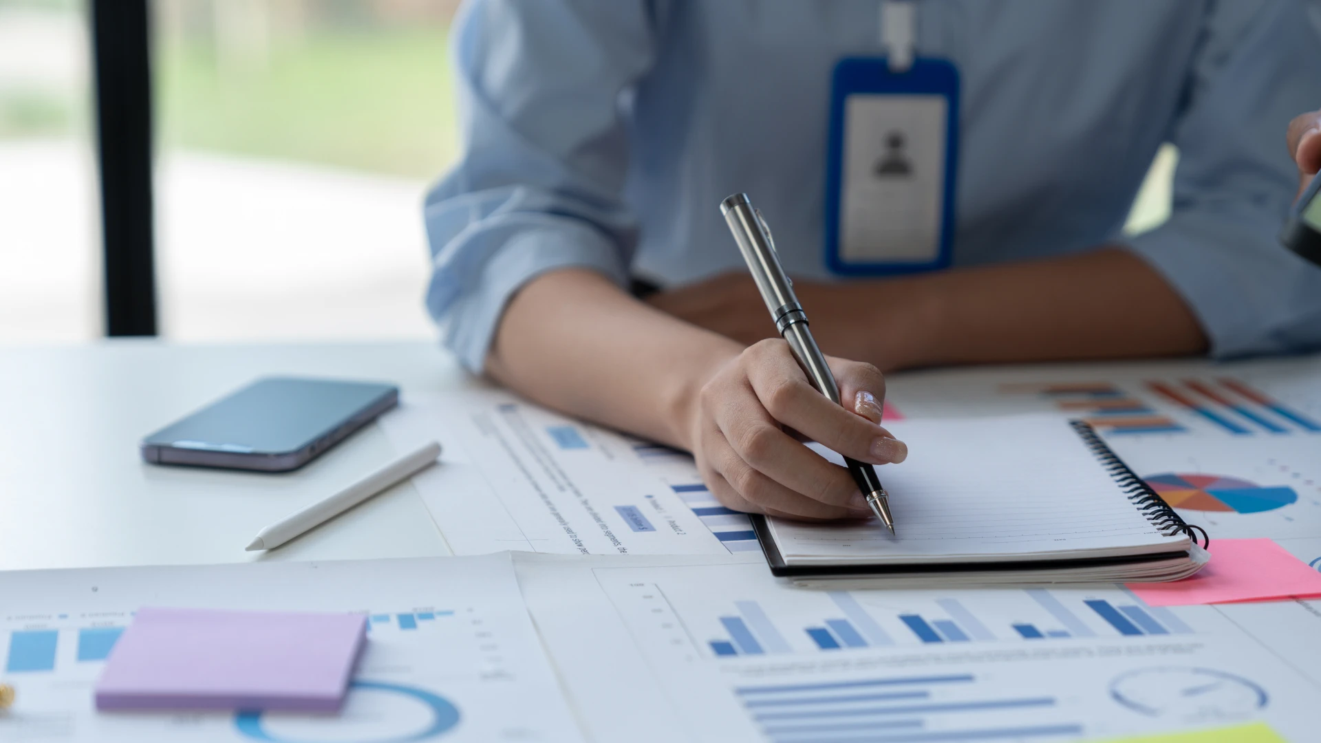 Person in a blue shirt taking notes, using a pen and spiral notebook.