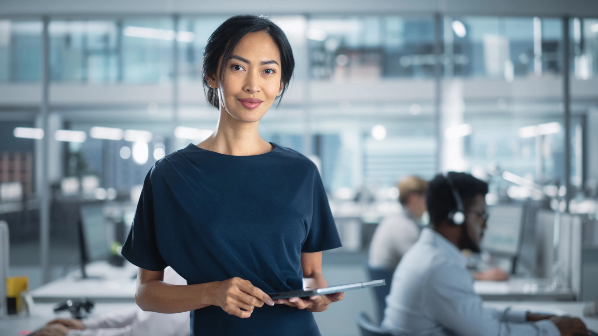 A woman in a blue shirt, standing in a modern office with employees working at computers.