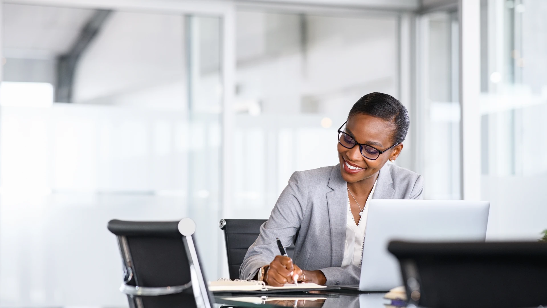 Smiling businesswoman with short hair and glasses working on a laptop in a bright, modern office.
