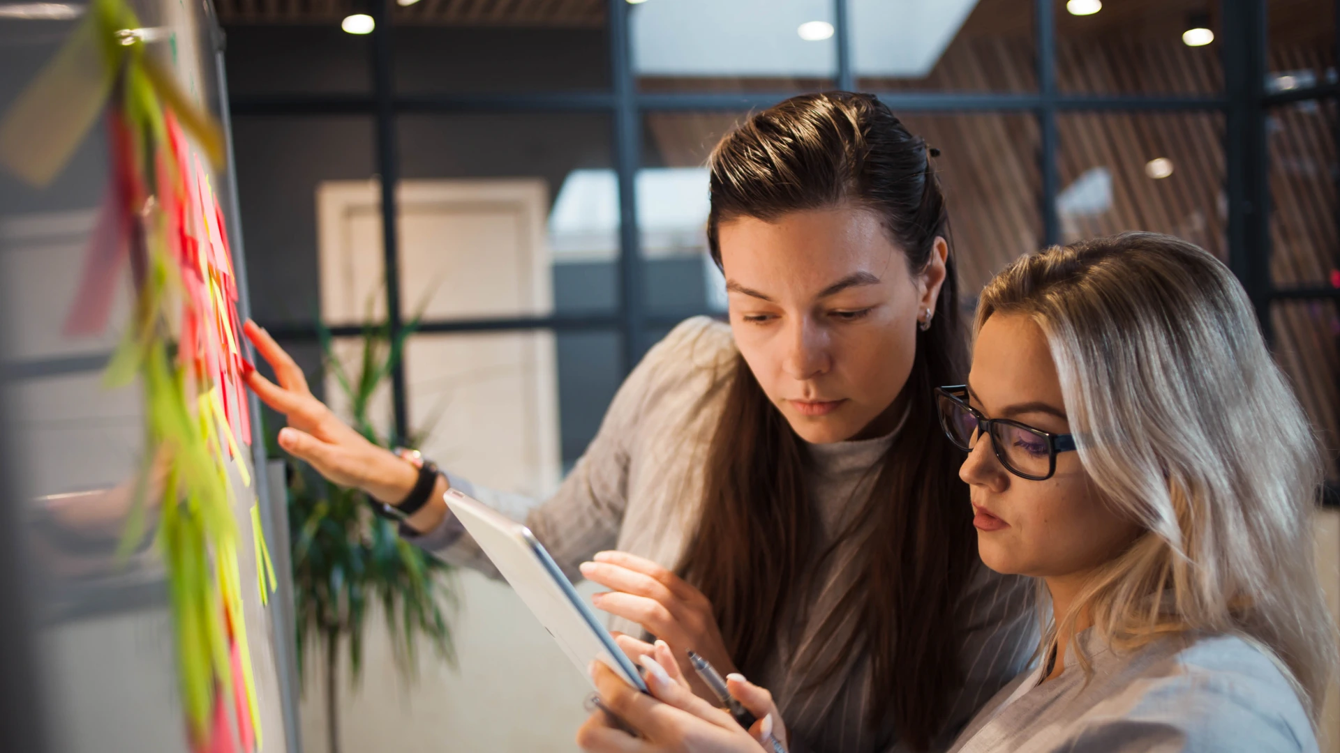 Two women are having a discussion. One is pointing at sticky notes on a board, while the other is holding a tablet and a pen.